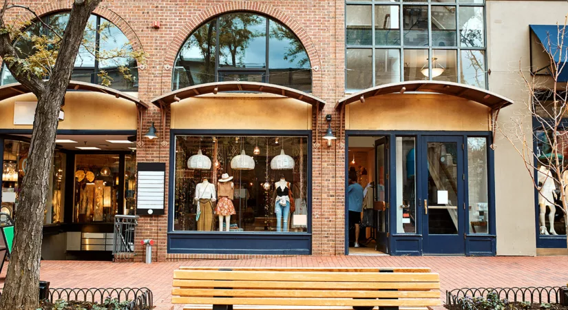 The exterior of a row of shops and retail spaces with brick walls, big windows, and a bench out front.