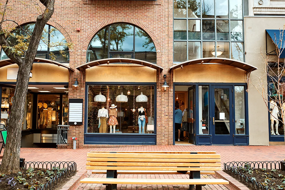 The exterior of a row of shops and retail spaces with brick walls, big windows, and a bench out front.